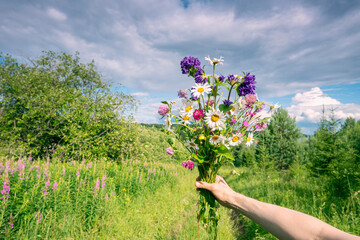 Wall Mural - a bouquet of wild flowers in your hand on the background of the river and the sky with clouds.