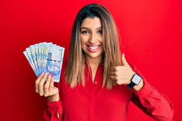 Poster - Beautiful brunette woman holding south african 100 rand banknotes smiling happy and positive, thumb up doing excellent and approval sign