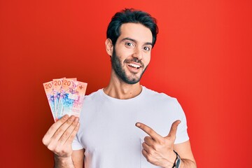 Sticker - Young hispanic man holding swiss franc banknotes smiling happy pointing with hand and finger