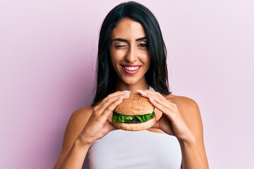 Wall Mural - Beautiful hispanic woman eating a tasty classic burger winking looking at the camera with sexy expression, cheerful and happy face.