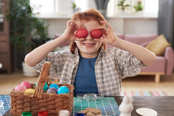 Wall Mural - Portrait of boy holding Easter eggs in front of his eyes and smiling at camera while sitting at the table with basket of eggs