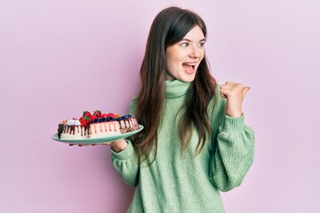 Sticker - Young beautiful caucasian girl holding cake slices pointing thumb up to the side smiling happy with open mouth