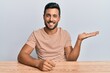 © Krakenimages.com - Handsome hispanic man wearing casual clothes sitting on the table smiling cheerful presenting and pointing with palm of hand looking at the camera.