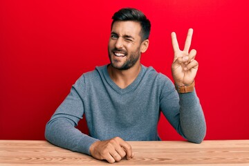 Poster - Handsome hispanic man wearing casual style sitting on the table smiling with happy face winking at the camera doing victory sign. number two.