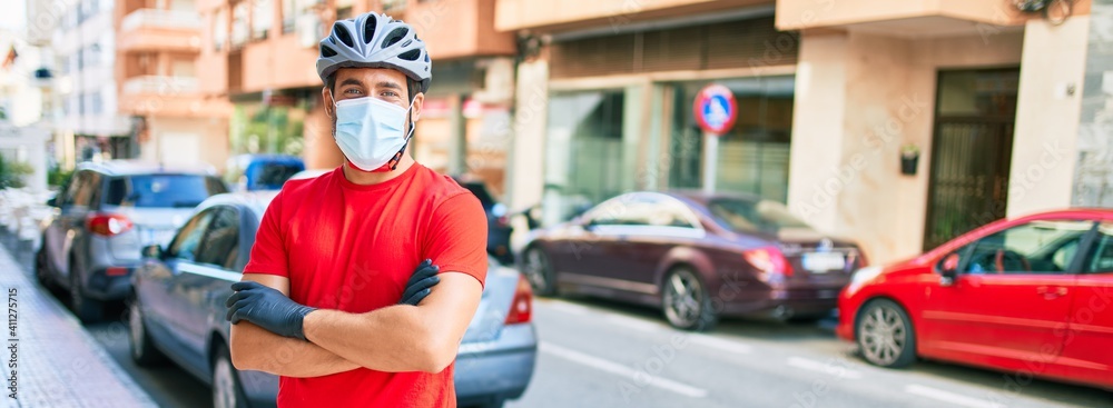 Young delivery man wearing bike helmet and coronavirus protection medical mask standing at town street. - obrazy, fototapety, plakaty 