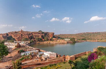 Wall Mural - Badami, Karnataka, India - November 7, 2013: Agasthya Lake and surrounding red rock hills. Some brown stone temple buildings sprinkled around. Green foliage and pink flowers add color.