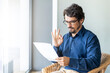 © Joao - Man wearing glasses on a chair holding paper document. Male portrait reading contract files and doing approved ok gesture at home office by the window