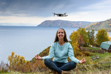 Wall Mural - Beautiful young woman having fun with a mini drone outdoors in Lotus pose. The girl sits against the autumn forest background