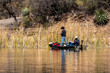 Two people fish from a small boat at Patagonia Lake State Park, Arizona. Both men are wearing face coverings. Their boat is overfilled with gear. Concepts of outdoor recreation, fishing, state parks