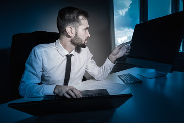 Poster - Office worker works late. Man next to a computer and laptop. Concept - overtime work. Office worker overtime work. Man in a white shirt works at night. Guy uses a computer and laptop at same time