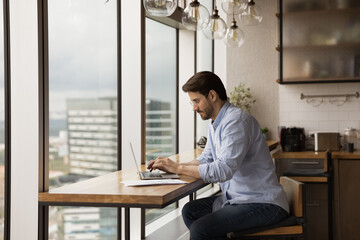 Concentrated young man in glasses renter owner of modern designed studio flat using laptop. Busy male manager working from home focused on computer screen typing on keyboard preparing documents online