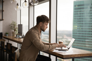 Concentrated millennial man write message on laptop keyboard at desk near panoramic window with splendid cityscape. Side shot of attentive young guy working on project document at modern office online