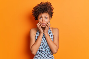 Wall Mural - Nervous worried Afro American woman clenches teeth looks embarrassed at camera wears fashiobnable outfit afraids of bad consquences poses in studio against orange background. Scared female model