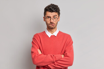 Wall Mural - Displeased Caucasian man bites lips keeps hands crossed dissatisfied with something wears round spectacles and sweater looks offended at camera poses against grey background. Negative emotions