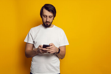 man with a beard and a white T-shirt stands with a phone on a yellow background
