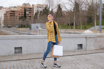 Wall Mural - Handsome young man wearing make up, laughing and speaking on his smartphone, carrying shopping bags. Non binary androgynous guy.