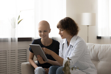 Wall Mural - Caring doctor in white medical uniform and young Caucasian bald female cancer patient look at tablet screen discuss good results. Happy GP and smiling woman with oncology excited with recovery.