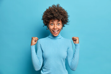 Excited successful Afro American woman clenches fists celebrates success in career says yes smiles broadly wears turtleneck isolated over blue background. Monochrome shot. Body language concept