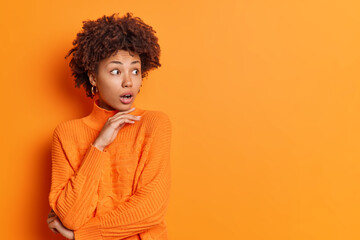 Wall Mural - Studio shot of stupefied African American woman holds chin and stares aside dressed in casual orange jumper reacts on astonishing relevation poses indoor blank copy space for your advertisement