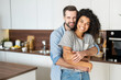 © Vadim Pastuh - Sweet multi-ethnic couple in love stands in embraces in the kitchen. Cheerful an African woman and a caucasian guy look at the camera and smile toothy