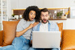 © Vadim Pastuh - Shocked multiracial couple looks at the laptop screen with opened mouths, an African woman and caucasian guy surprised with unexpected new, watching a scary movie sitting on the couch at home