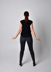 Canvas Print - Full length portrait of woman with red hair in a ponytail, wearing black t-shirt and denim pants. Standing pose, hands reaching out with back to the camera the camera, against a  studio background.