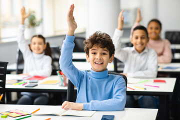 Diverse small schoolkids raising hands at classroom