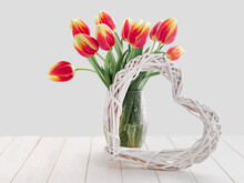 Bunch Of Red Yellow Stripe Tulips In Glass Vase. Whitewashed Wattle Rattan Wreath In Shape Of Heart. Spring Decorations On Off White, Natural Rustic Wooden Table.