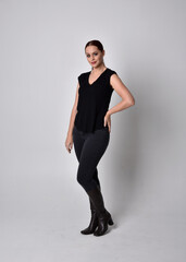 Poster - Simple full length portrait of woman with red hair in a ponytail, wearing casual black tshirt and jeans. Standing pose facing front on, against a  studio background.