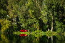 Red Camper Van On Shore Of Danube River With Green Trees And Grass In Background And With Reflecion In Water Surface.