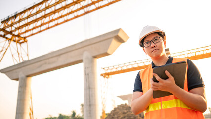 Smart Asian worker man or male civil engineer with protective safety helmet and reflective vest using digital tablet for project planning and checking architectural drawing at construction site.