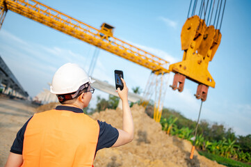 Wall Mural - Smart Asian worker man or male civil engineer with protective safety helmet and reflective vest using using smartphone for taking photo at construction site.