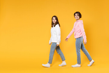 Wall Mural - Full length side view of cheerful smiling funny two young women friends 20s wearing casual white pink hoodies walking going looking camera isolated on bright yellow color background studio portrait.