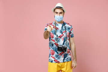 Wall Mural - Funny traveler tourist man in face mask to safe from coronavirus virus covid-19 point index finger camera on youn isolated on pink background. Passenger travel on weekend. Air flight journey concept.