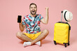 Full length happy traveler tourist man sit on floor hold mobile phone with blank empty screen doing winner gesture isolated on pink background. Passenger travel on weekend. Air flight journey concept.