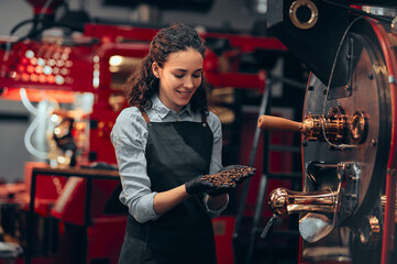 Woman checking the quality of the coffee beans standing near the roaster machine at the roastery. Mixing roasted coffee.