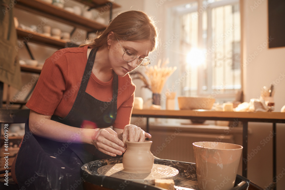 Warm toned portrait of young female artisan shaping clay on pottery wheel in sunlit workshop and enjoying arts and crafts, copy space - obrazy, fototapety, plakaty 