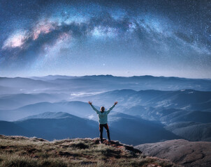 Wall Mural - happy man on the mountain peak and arched milky way over mountains in low clouds at night. landscape