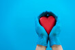 Hands wearing blue medical gloves holding red heart over light blue background. Health care, heart disease, cardiology concept