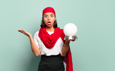 Wall Mural - young latin woman looking surprised and shocked, with jaw dropped holding an object with an open hand on the side