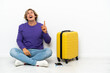 © luismolinero - Young blonde man with suitcase sitting on the floor pointing up and surprised