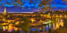 Panorama Cityscape Of Old Town Of Bern With Cathedral Tower And Nydeggbr Cke Bridge Illuminated At Night Reflecting Into Aare River. Popular Landmark Of Historical Town UNESCO World Heritage Site.