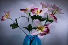 A Pretty Display Of Pink And Red Artificial Flowers In A Translucent Blue Vase Isolated On A Pale Blue Background.