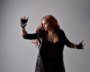 Sticker - Close up portrait of girl with long red hair wearing dark leather coat, corset and gloves. Posing with gestural hand movements as if casting spell. Moody back lit lighting against a studio background.