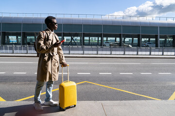 Afro-American millennial traveler man with suitcase stands in airport terminal, holding cellphone, calling and looking for a taxi or car sharing. Black male waiting for cab for transporting