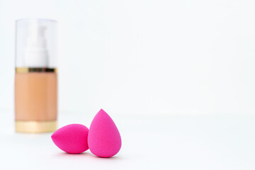 Wall Mural - Close-up of clean pink sponge on the background of a bottle of beige cosmetic foundation for make up. Makeup artist accessories, products for perfect face skin on a white table. Copy space.