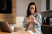 Happy charming woman using smartphone while working with laptop