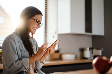 Wall Mural - Happy charming woman smiling while using smartphone at home kitchen