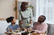 © Seventyfour - Portrait of modern African-American woman serving food for family while enjoying dinner together in cozy home interior, copy space