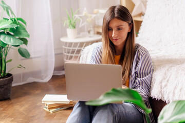 Distance learning online education and work. Business woman typing at laptop, sitting on the floor near the plants and books. Smiling girl with notebook at home office. Using computer and shopping.